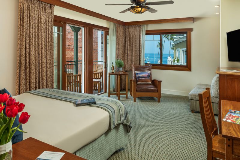 A spacious hotel suite featuring a king-sized bed with a blue-striped throw, a wooden desk and chair in the foreground, and a large sliding glass door that opens to a balcony with a scenic view of the ocean.