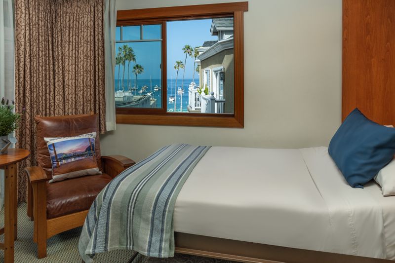 A view from a hotel window shows a cozy brown leather armchair with a decorative pillow, a sofa bed with a blue-and-white striped throw, and a window looking out toward a sunny harbor with palm trees and the ocean in the distance.