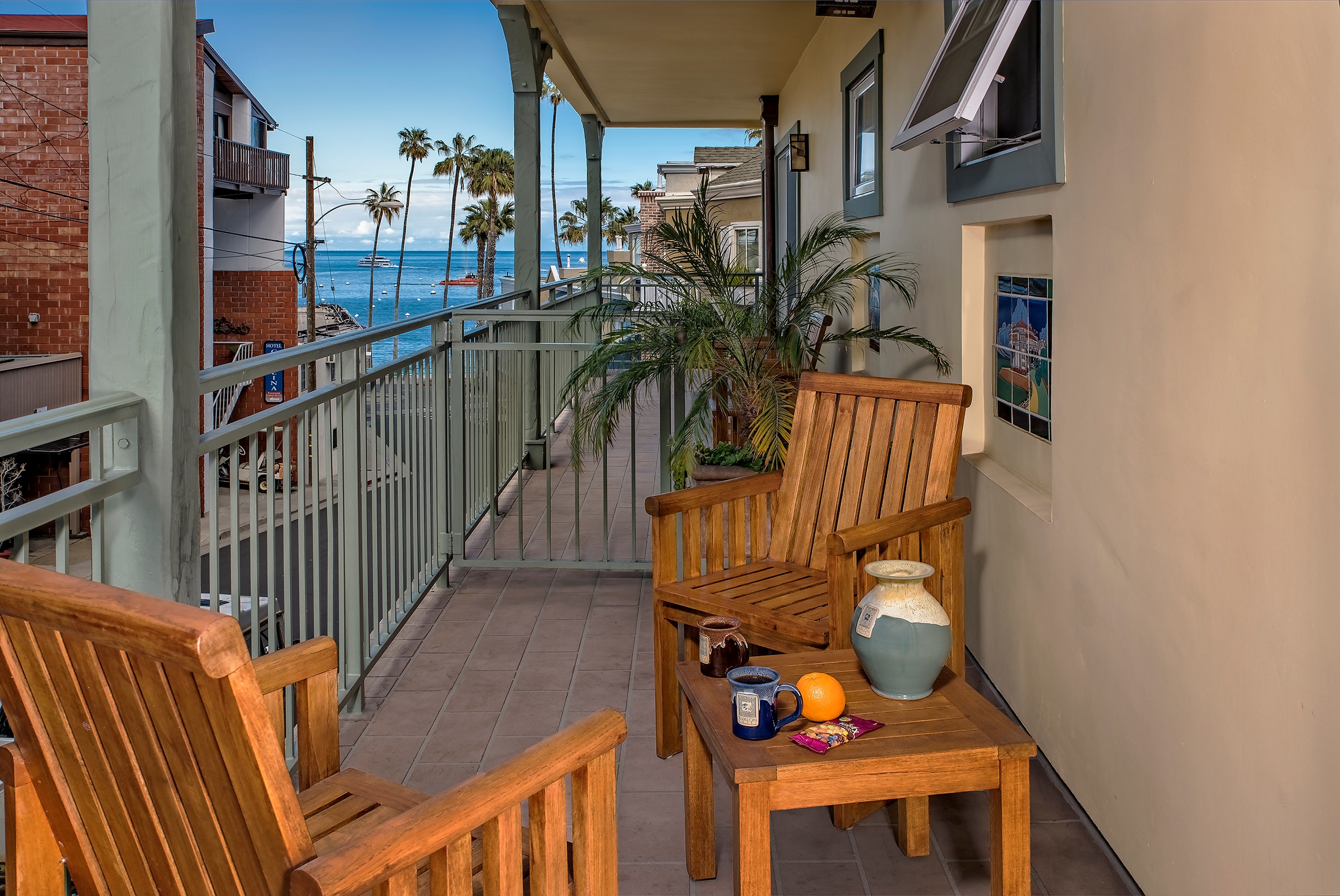 A long outdoor balcony with wooden chairs and a small table set with a blue mug and fruit, offering a view of a street lined with palm trees leading toward the blue ocean.
