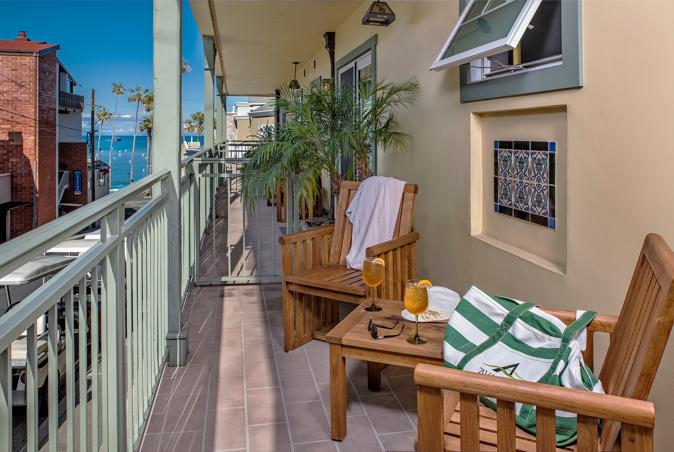 A narrow, sun-drenched, covered balcony with wooden chairs and a small table featuring drinks, overlooking a coastal town with palm trees and a glimpse of the blue ocean in the distance.
