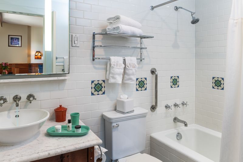 A bright, white-tiled bathroom featuring a white marble vanity with a vessel sink and Fiesta ware accessories, a combination bathtub and shower, and decorative green and white Catalina tiles on the walls.
