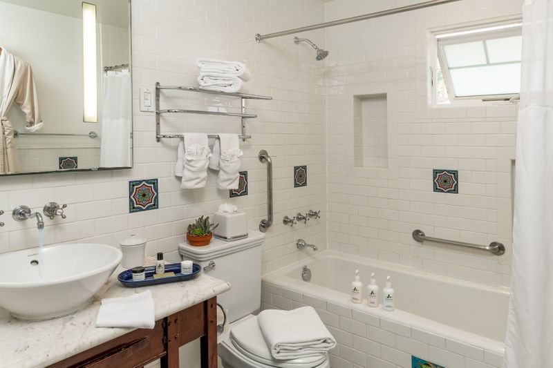 A clean hotel bathroom featuring a white marble vanity with a vessel sink, a combination bathtub and shower with silver fixtures, and white tiled walls accented by decorative square Catalina tiles with blue and green floral patterns.