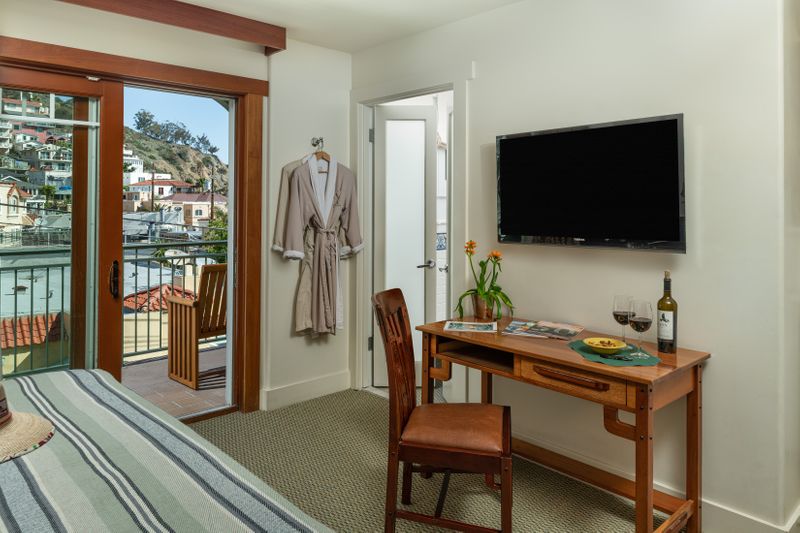 A bright hotel room featuring a wooden writing desk with a chair and a vase of sunflowers, a wall-mounted flat-screen TV, and an open sliding door leading to a balcony with lush greenery.