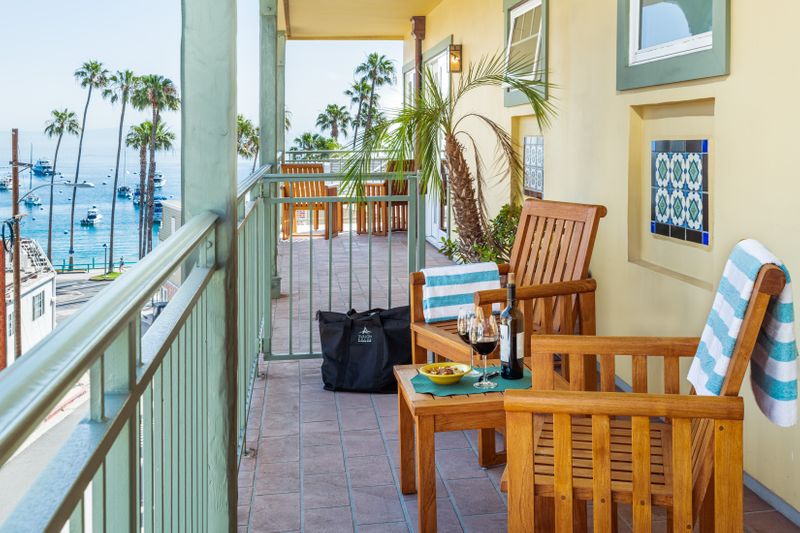 A long outdoor balcony with wooden chairs and a small table set with a wine bottle and snacks, overlooking a scenic coastal view of the ocean and palm trees under a bright blue sky.