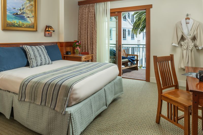 A bright hotel bedroom featuring a bed with navy blue and striped pillows, a wooden desk with a chair, and a sliding glass door that opens to a balcony with a view of a palm tree.