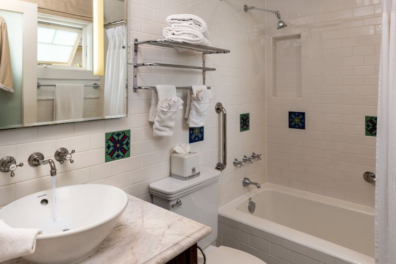 A clean, white-tiled bathroom featuring a white marble vanity with a vessel sink, a combination bathtub and shower with silver fixtures, and decorative blue and green Catalina tiles.