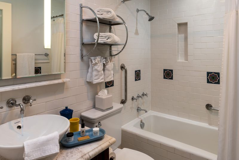 A clean, white-tiled bathroom featuring a marble vanity with Fiesta ware accessories and a vessel sink, a combination bathtub and shower with silver fixtures, and decorative Catalina tiles.