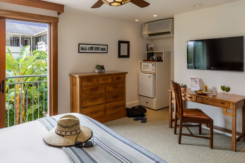 A hotel room interior featuring a king bed, a wooden dresser and a matching desk with a chair, a wall-mounted TV, and an open sliding glass door that leads to a balcony with lush green foliage.