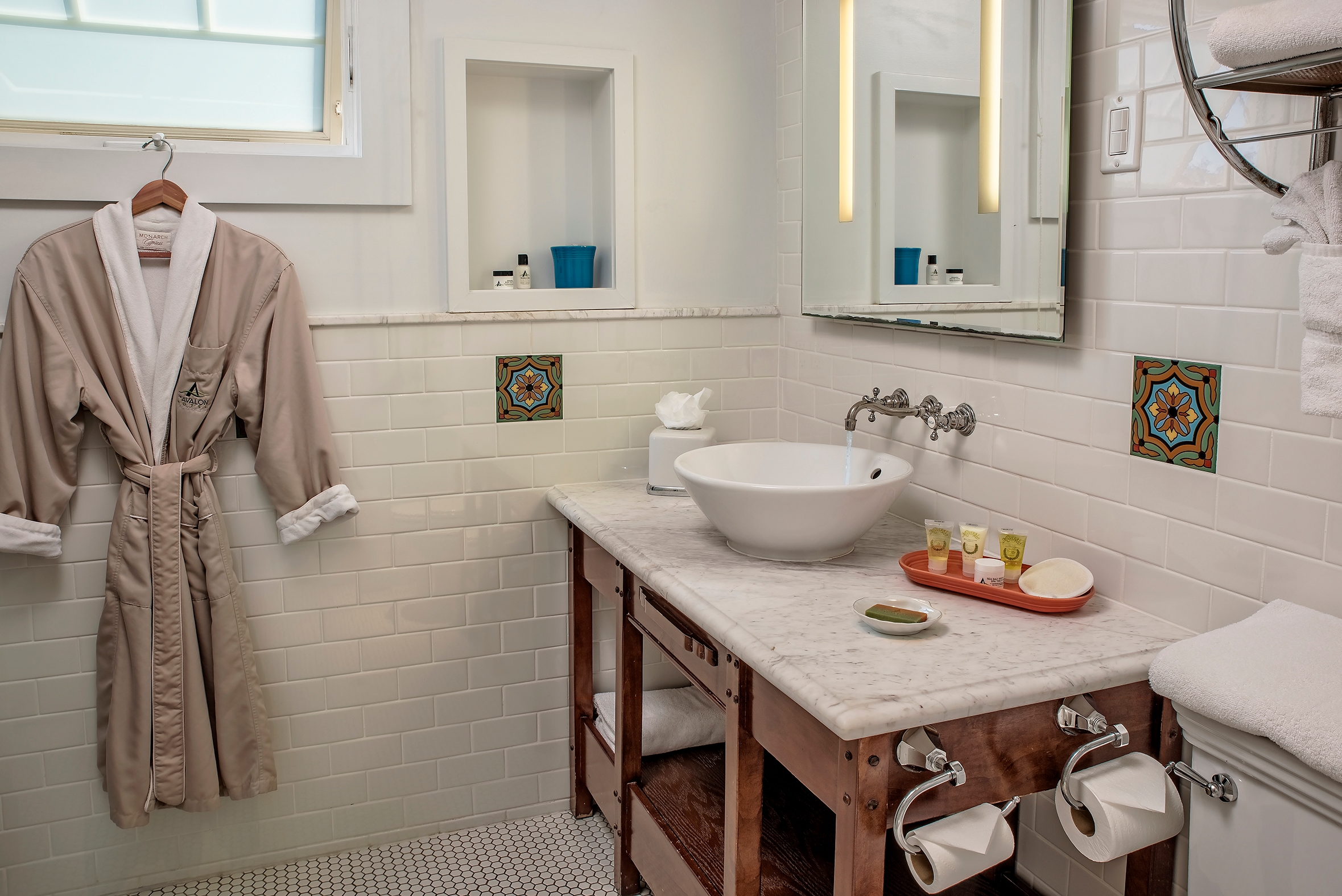 A hotel bathroom with white subway tile walls featuring a marble-topped wooden vanity with a vessel sink, a large mirror with integrated lighting, and a tan bathrobe hanging on the wall.