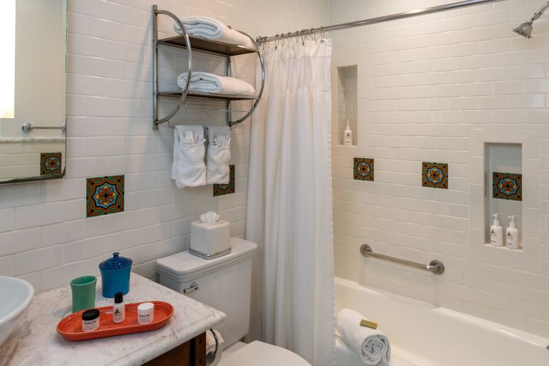 A clean hotel bathroom featuring white subway tile walls, a marble-topped wooden vanity with a vessel sink and Fiesta ware accessories, and a large mirror with integrated side lighting. A tan bathrobe hangs on the wall next to a window, and the space is accented with decorative patterned tiles.