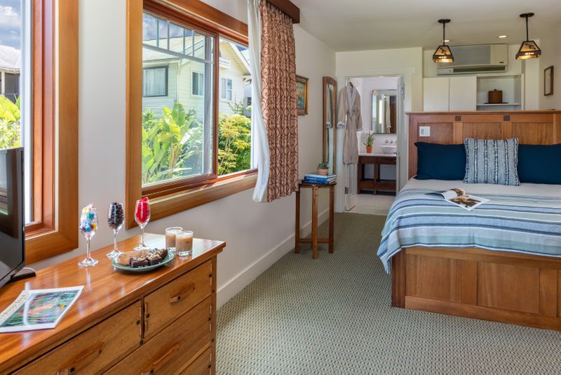 A bright hotel room featuring a bed with a blue-and-white striped throw, a wooden dresser in the foreground with wine glasses and snacks, and large windows that offer a view of lush green foliage and neighboring coastal buildings.