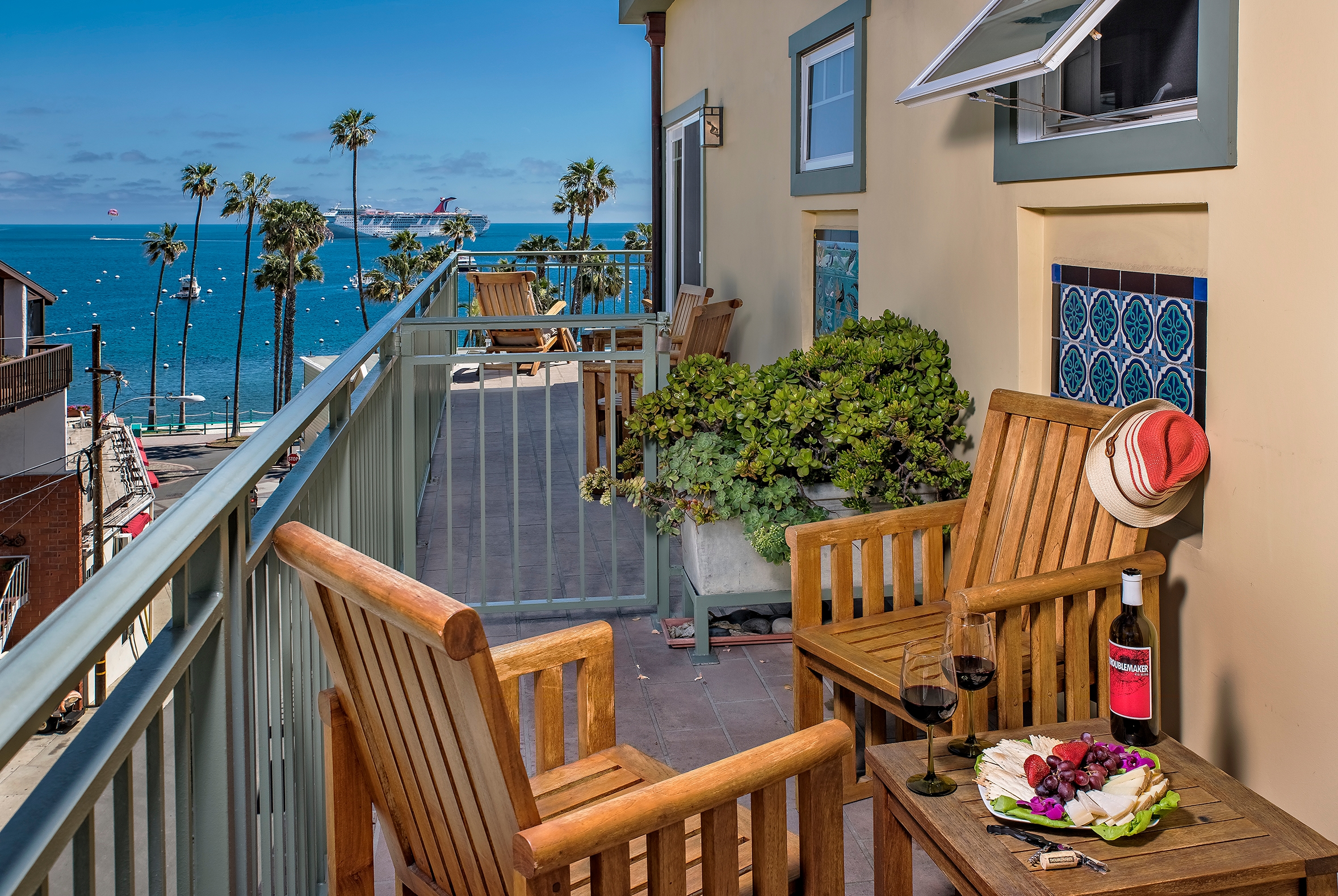 A long, narrow outdoor balcony with wooden chairs and a small table set with wine and a platter of fruit and cheese, overlooking a scenic coastal view of the ocean and palm trees under a bright blue sky.