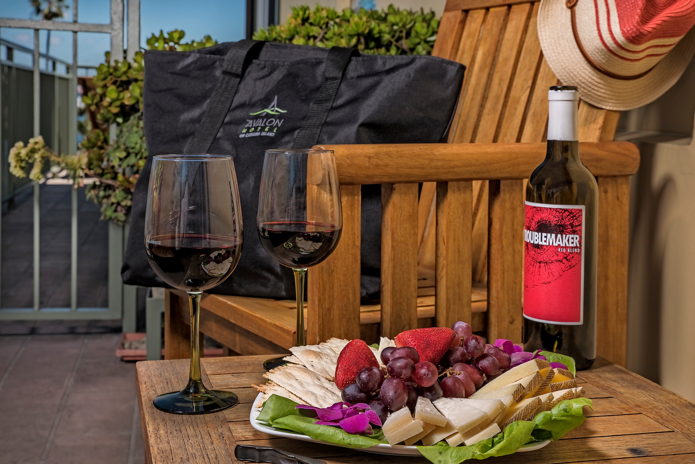A close-up of a wooden outdoor table on a balcony featuring two glasses of red wine, a bottle of wine, and a white platter filled with grapes, strawberries, cheese, and crackers.