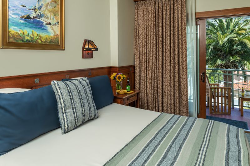 A close-up view of a hotel bed with a white duvet and striped throw, navy blue pillows, and a wooden headboard, with a sliding glass door in the background opening to a balcony with a palm tree.
