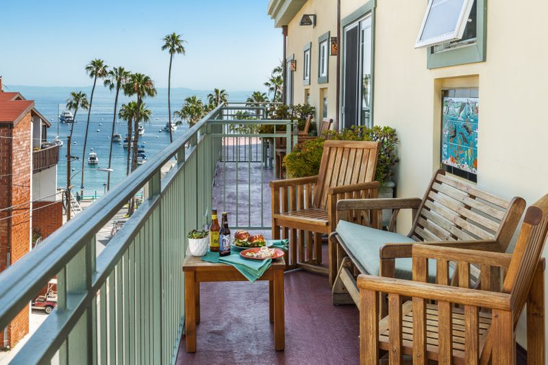 A long, narrow outdoor balcony with wooden chairs and a small table set with snacks and drinks, offering a picturesque view of a coastal town, palm trees, and the ocean under a clear blue sky.