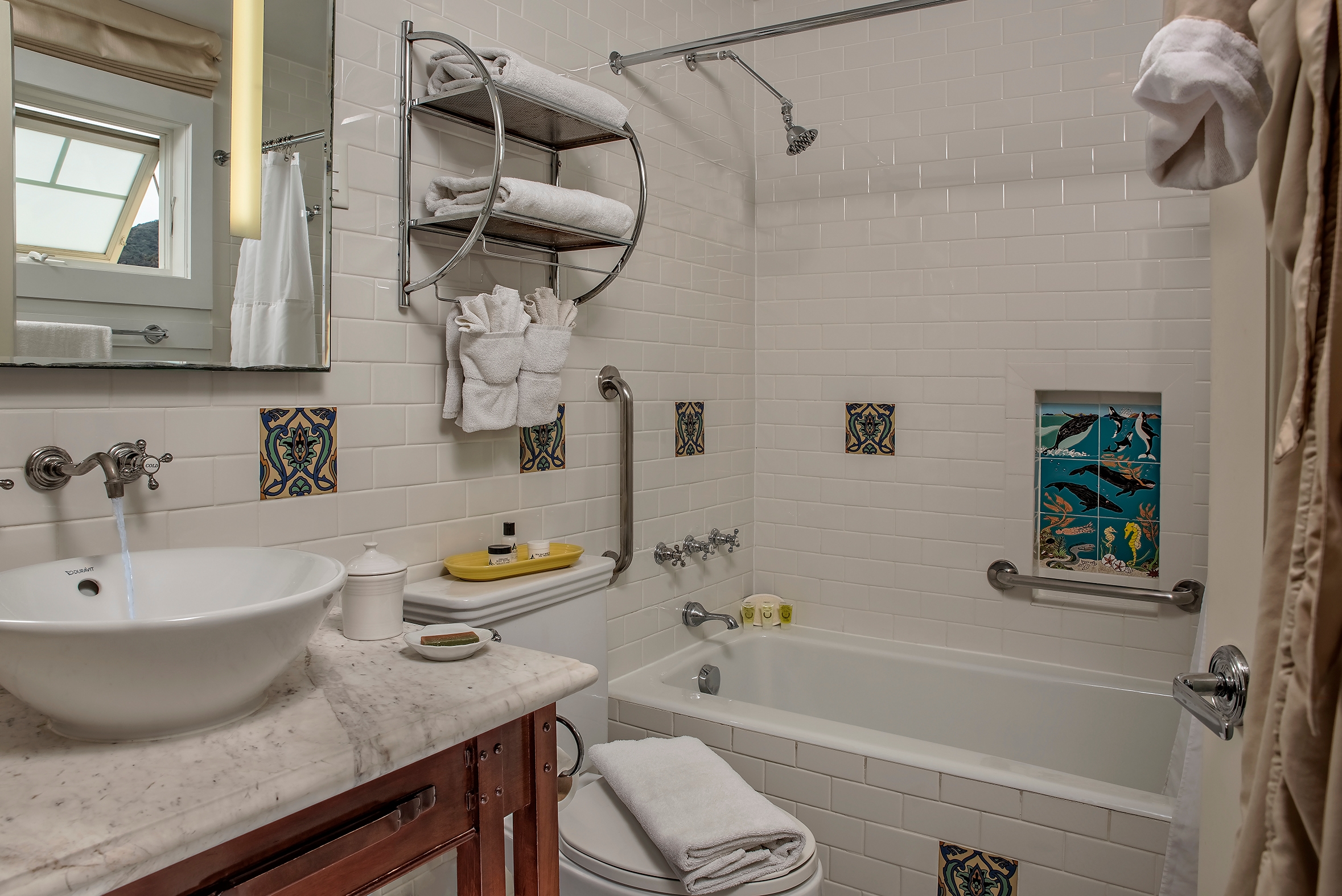 A clean, white-tiled bathroom featuring a white marble vanity with a vessel sink, a combination bathtub and shower with silver fixtures, and decorative blue and yellow tiles.