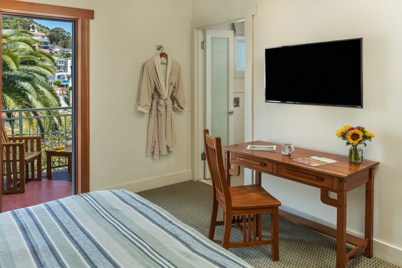 A bright hotel room featuring a wooden writing desk with a chair and a vase of sunflowers, a wall-mounted flat-screen TV, and an open sliding door leading to a balcony with lush greenery.