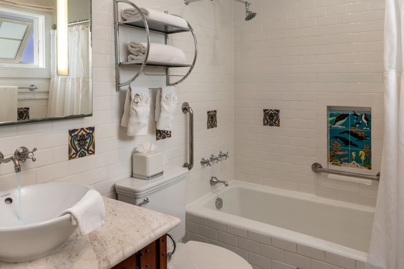 A bright hotel bathroom featuring a white marble vanity with a vessel sink, a combination bathtub and shower with silver fixtures, and decorative square Catalina tiles with various blue and green floral patterns.