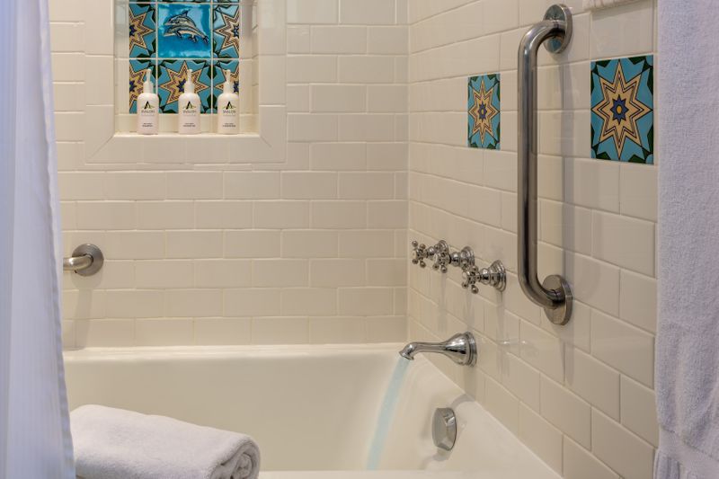 A clean hotel bathroom featuring white subway tile walls, a marble-topped wooden vanity with a vessel sink, and a large mirror with integrated side lighting. A tan bathrobe hangs on the wall next to a window, and the space is accented with decorative patterned tiles.