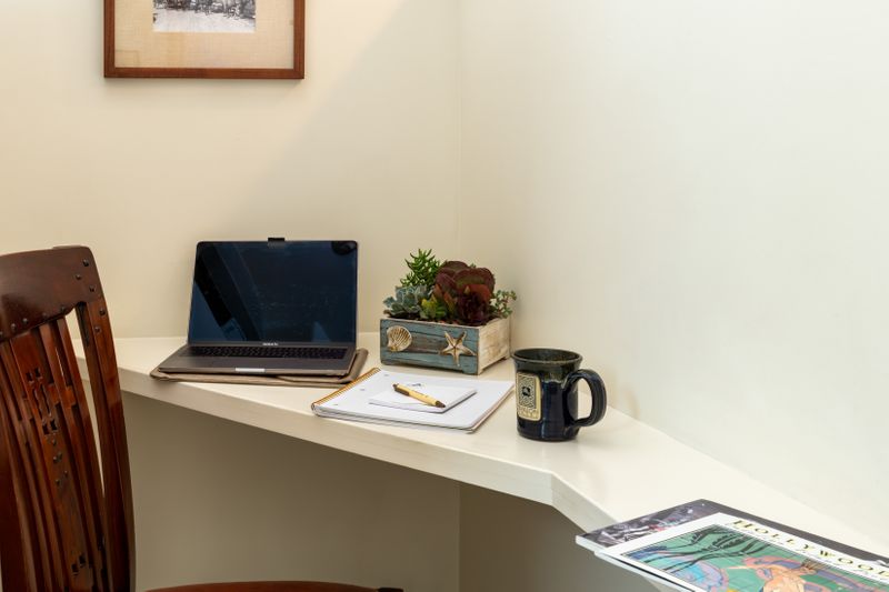 A minimalist in-room workspace featuring a curved white built-in desk, a dark wood chair, a laptop, a notebook with a pen, a small succulent planter, and a black mug.