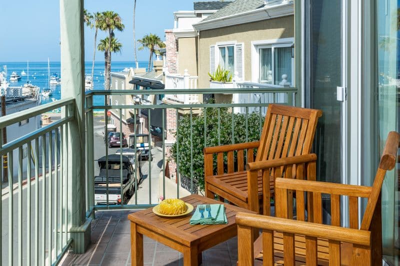 An outdoor balcony with wooden armchairs and a small table overlooking a coastal town and harbor, featuring a view of the ocean, palm trees, and neighboring buildings under a clear blue sky.
