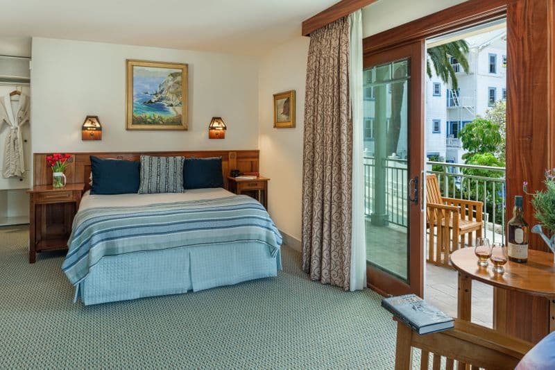 A cozy hotel bedroom featuring a king-sized bed with a blue-and-white striped throw, a wooden headboard with built-in lamps, and a view of a scenic hillside town through an open sliding glass door.