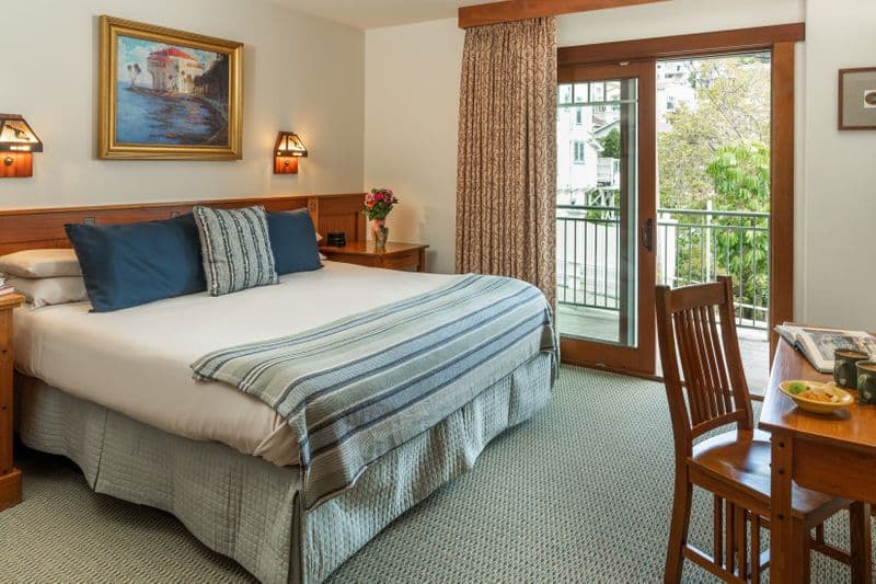 A bright hotel room with a neatly made bed featuring a blue and white striped throw, a wooden writing desk with a chair, and a sliding glass door that leads to a sunlit balcony overlooking lush greenery.