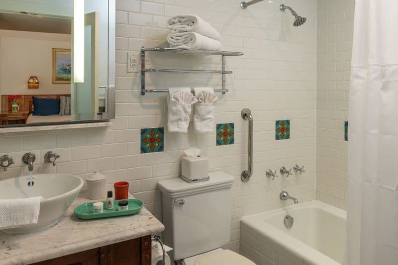 A clean, white-tiled bathroom featuring a white marble vanity with a vessel sink, a combination bathtub and shower with silver fixtures, and decorative square tiles with green and brown patterns.
