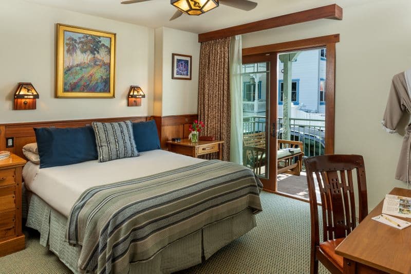 An inviting, traditional hotel room featuring a queen-sized bed with navy blue and striped pillows, and a sliding glass door that leads to a private balcony.