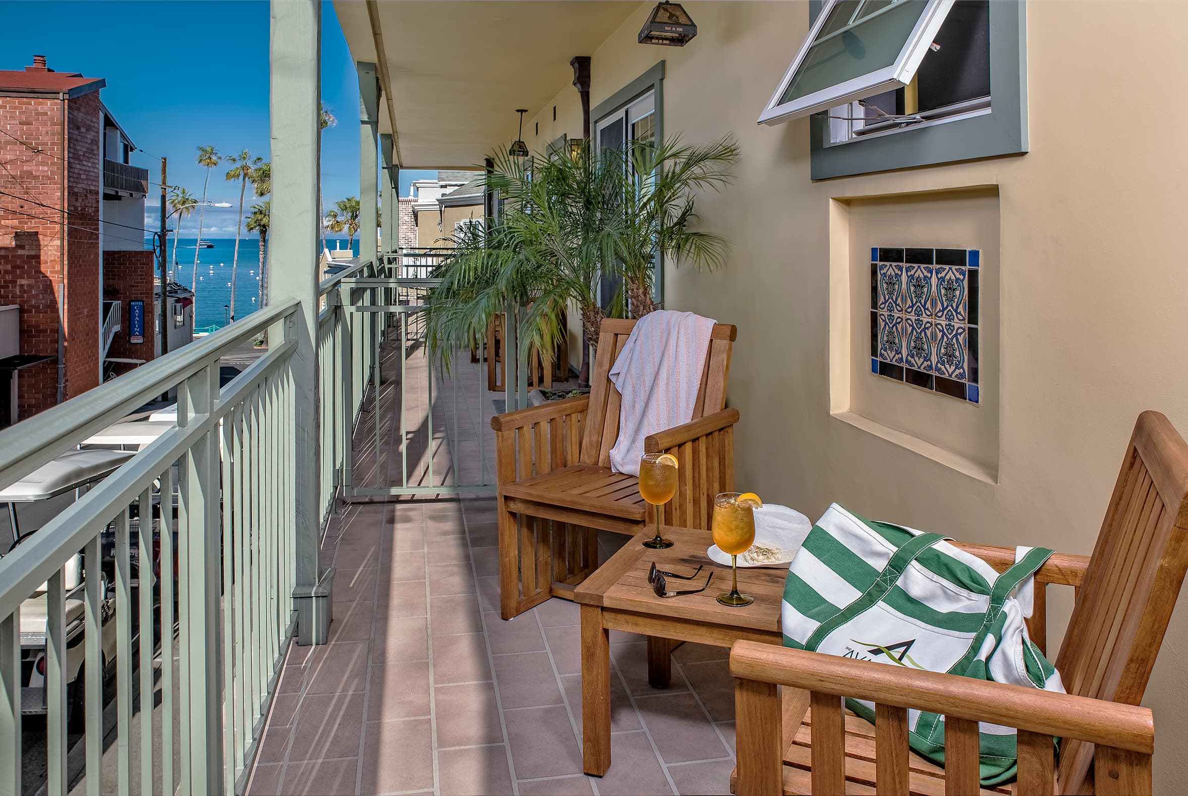 A narrow, sun-drenched, covered balcony with wooden chairs and a small table featuring drinks, overlooking a coastal town with palm trees and a glimpse of the blue ocean in the distance.