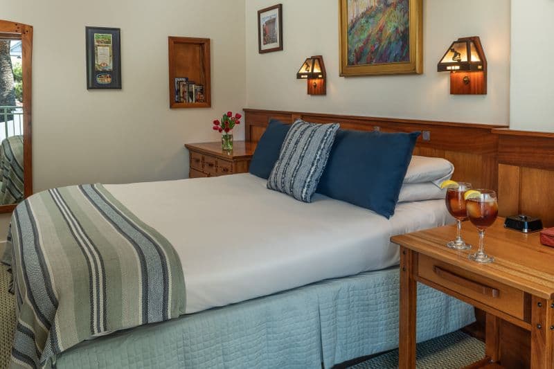 A cozy bedroom with a wooden headboard and matching nightstand, featuring a queen bed neatly made with a striped throw and navy blue pillows, accented by warm lighting from wall-mounted lamps.