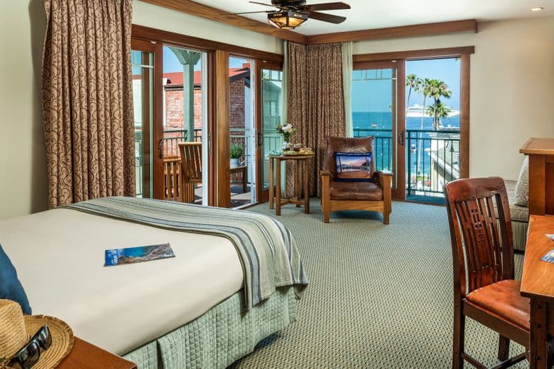 A bright hotel room featuring a bed with a blue-and-white striped throw, a brown leather armchair with a decorative pillow, and a large window and sliding glass door that reveal a scenic view of a sunny harbor with boats and palm trees.