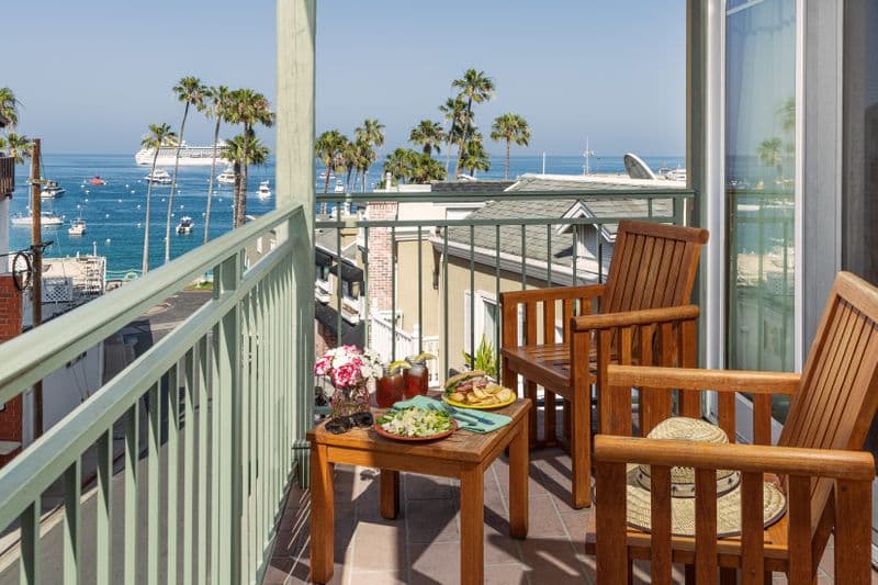 A private hotel balcony with two wooden armchairs and a small side table holding snacks, overlooking a vibrant blue harbor filled with boats and lined with tall palm trees under a clear sky.