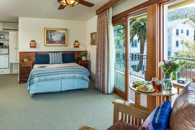 A bright hotel room featuring a queen bed with a blue-and-white striped throw, a wooden headboard, and an open sliding glass door leading to a balcony with a view of a large palm tree and a white building.