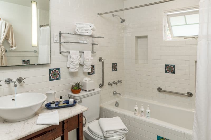 A clean hotel bathroom featuring a white marble vanity with a vessel sink, a combination bathtub and shower with silver fixtures, and white tiled walls accented by decorative square Catalina tiles with blue and green floral patterns.