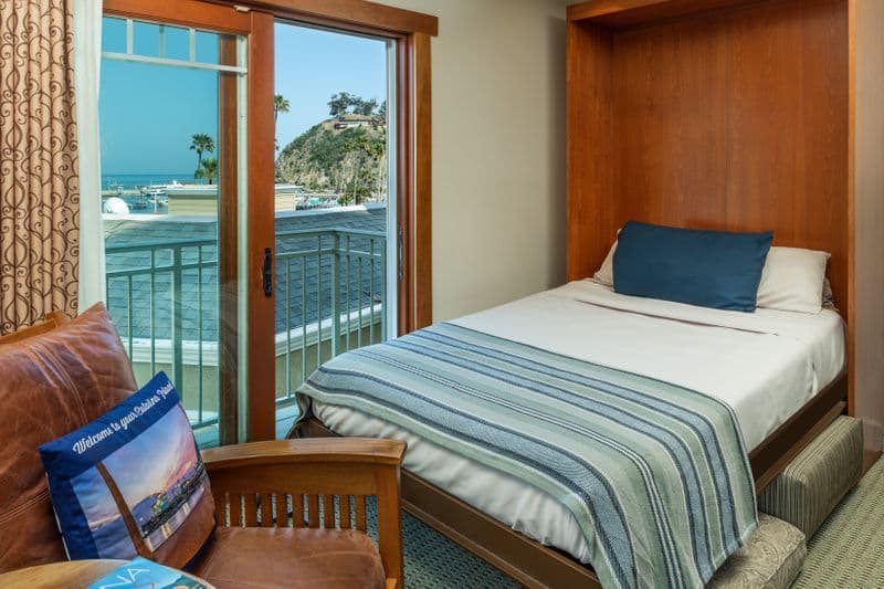 A brightly lit hotel room featuring a fold-down Murphy bed with a blue-striped throw, a brown leather armchair with a scenic decorative pillow, and an open sliding glass door that leads to a private balcony with a view of a vibrant blue harbor and tall palm trees.