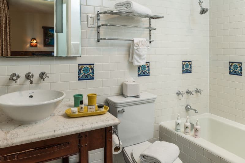 A bright, white-tiled bathroom featuring a white marble vanity with a vessel sink and Fiesta ware accessories, a combination bathtub and shower with silver fixtures, and decorative blue and yellow Catalina tiles.