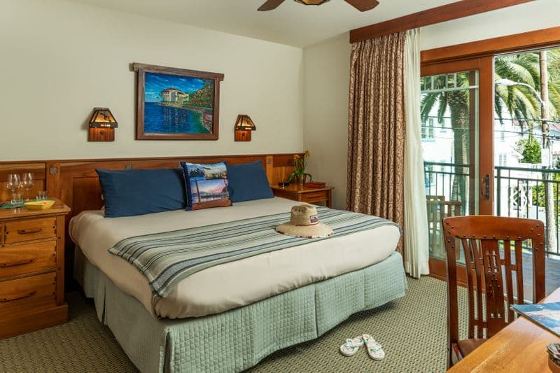 A bright hotel bedroom featuring a bed with navy blue and striped pillows, a wooden nightstand with a small vase of flowers, and a sliding glass door that leads to a sunlit balcony with a view of a palm tree.