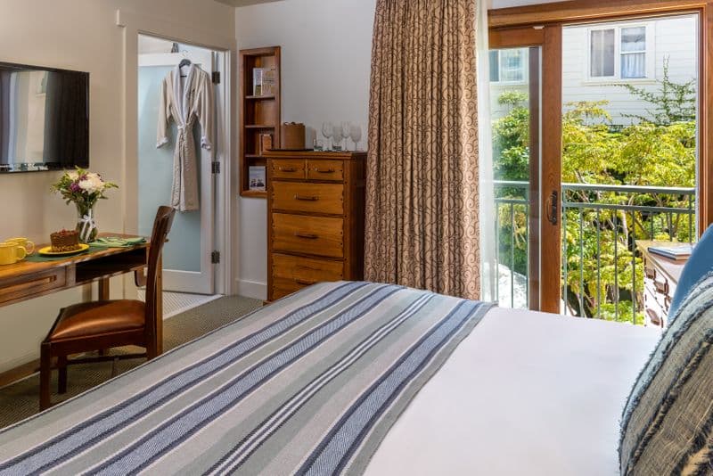 A bright hotel bedroom featuring a bed with navy blue and striped pillows, a wooden dresser, and a sliding glass door leading to a balcony with a view of lush greenery.