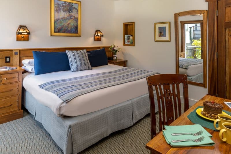 A bright hotel bedroom featuring a bed with navy blue and striped pillows, a wooden dresser, and a sliding glass door leading to a balcony with a view of lush greenery.