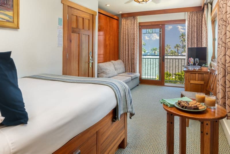 A bright hotel room featuring a bed with a blue-and-white striped throw, a brown leather armchair with a decorative pillow, and a large window and sliding glass door that reveal a scenic view of a sunny harbor with boats and palm trees.