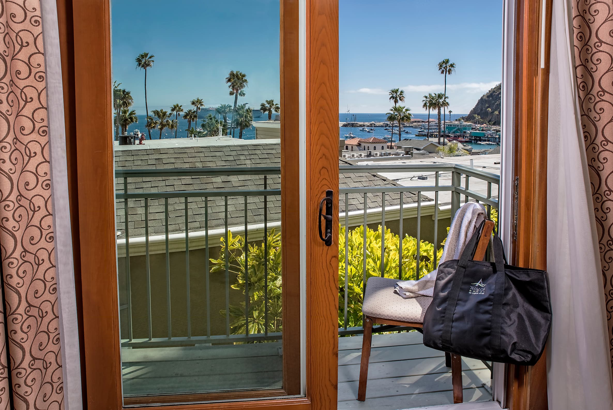 A private hotel balcony with a black travel bag resting on a chair, looking out through an open sliding glass door toward a sunny harbor with boats, tall palm trees, and a coastal hillside.