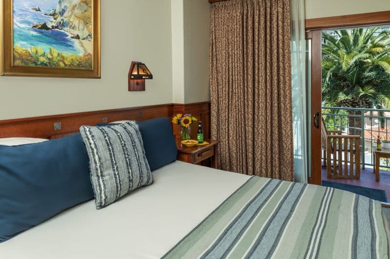 A close-up view of a hotel bed with a white duvet and striped throw, navy blue pillows, and a wooden headboard, with a sliding glass door in the background opening to a balcony with a palm tree.