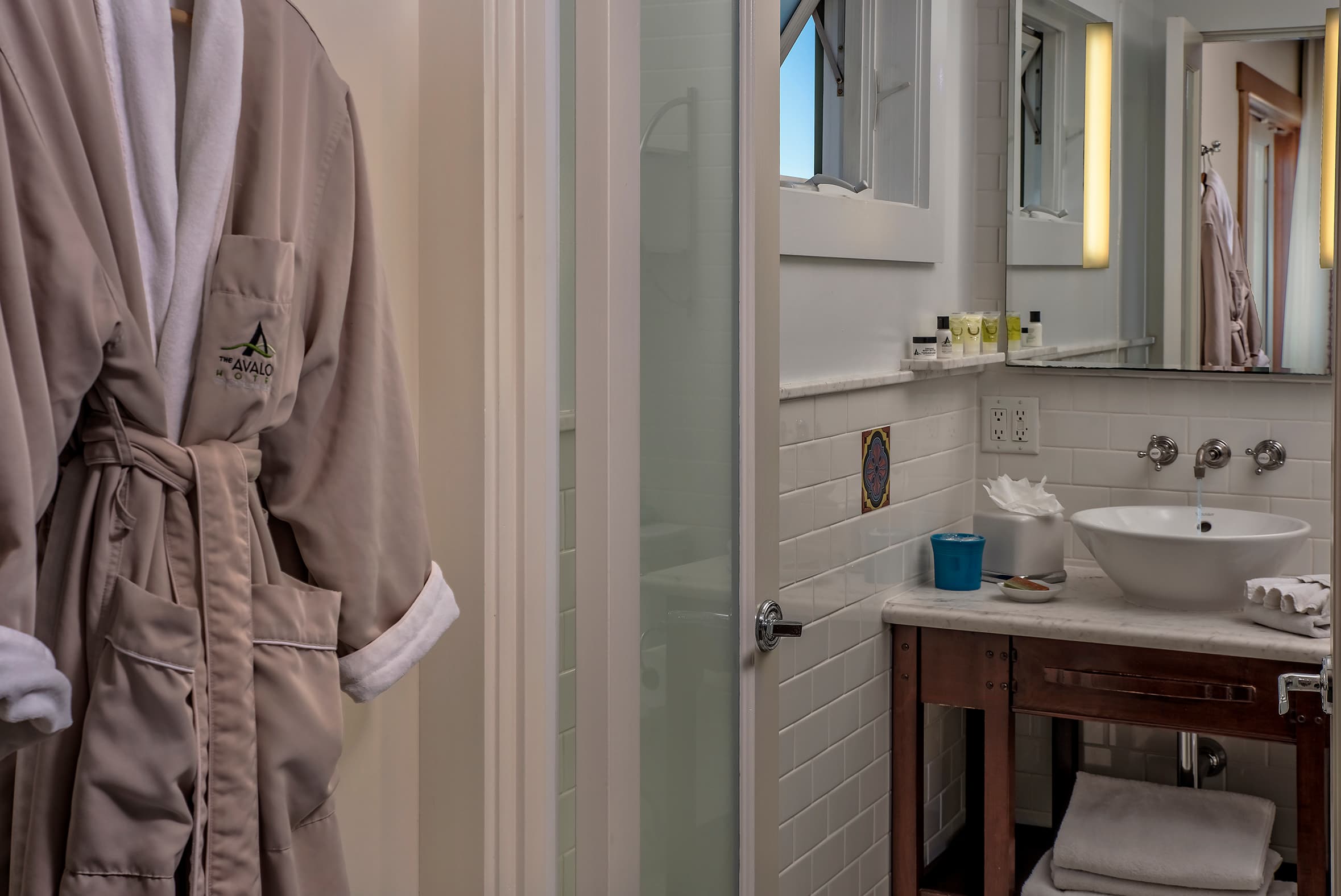 A plush tan bathrobe hangs on a white door in the foreground, with a partial view into a brightly lit bathroom featuring a marble vanity and a vessel sink in the background.