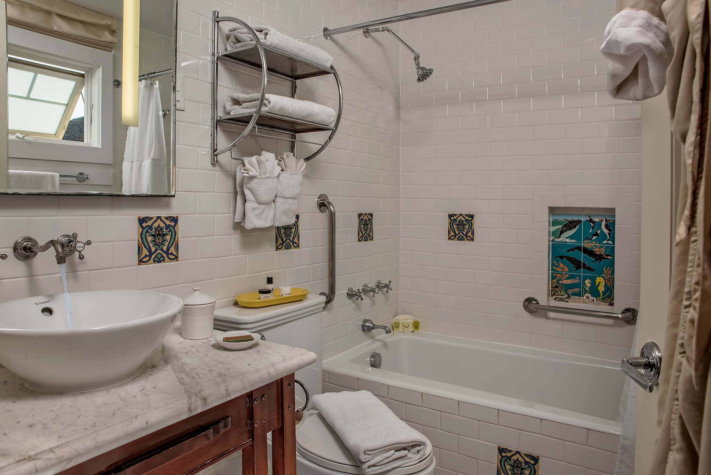 A clean, white-tiled bathroom featuring a white marble vanity with a vessel sink, a combination bathtub and shower with silver fixtures, and decorative blue and yellow tiles.