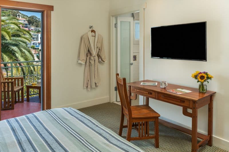 A bright hotel room featuring a wooden writing desk with a chair and a vase of sunflowers, a wall-mounted flat-screen TV, and an open sliding door leading to a balcony with lush greenery.