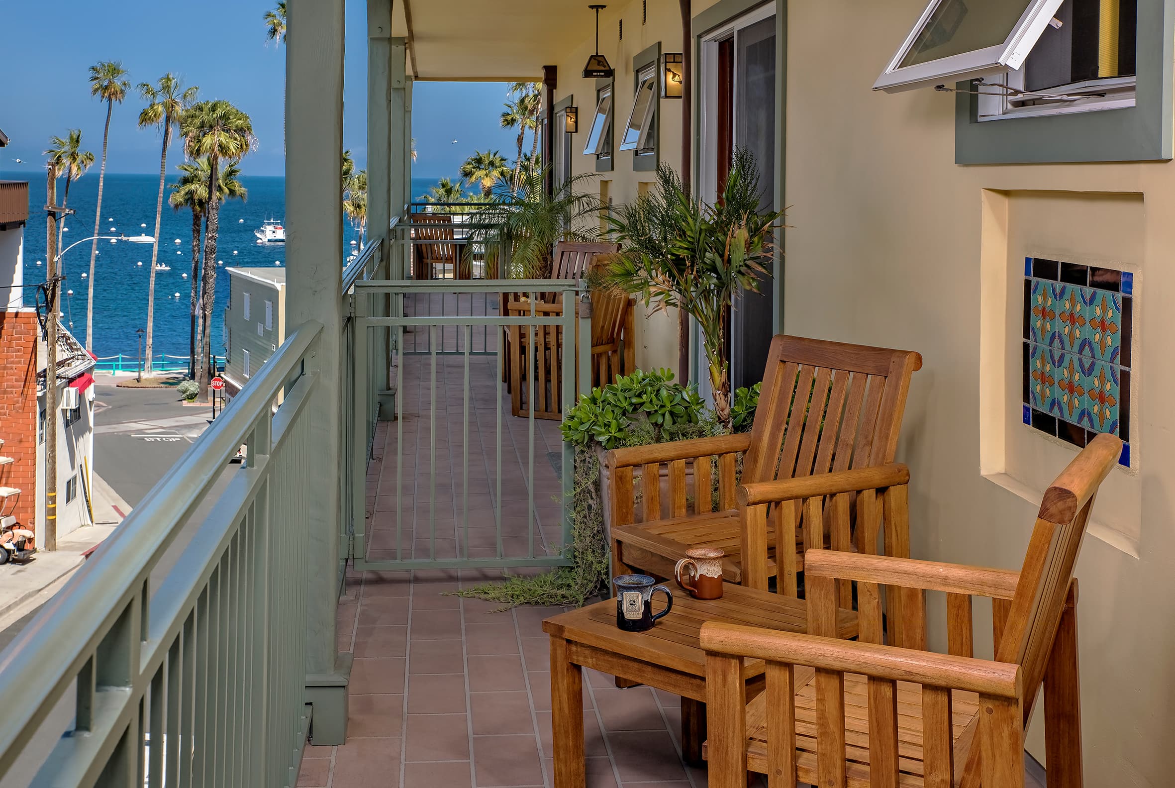 A long, narrow outdoor balcony with wooden chairs and a small table, featuring a view of a coastal street lined with palm trees leading toward the blue ocean under a clear sky.