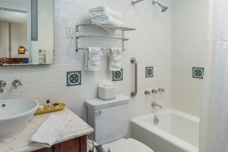 A clean, white-tiled bathroom featuring a white marble vanity with a vessel sink, a combined bathtub and shower with a silver grab bar, and decorative green patterned tiles on the wall.