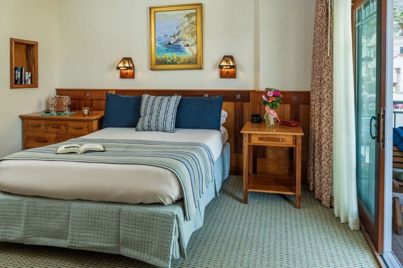 A bright hotel bedroom featuring a neatly made bed with navy blue and striped pillows, a wooden nightstand with a bouquet of flowers, and a sliding glass door that leads to a sunlit balcony.