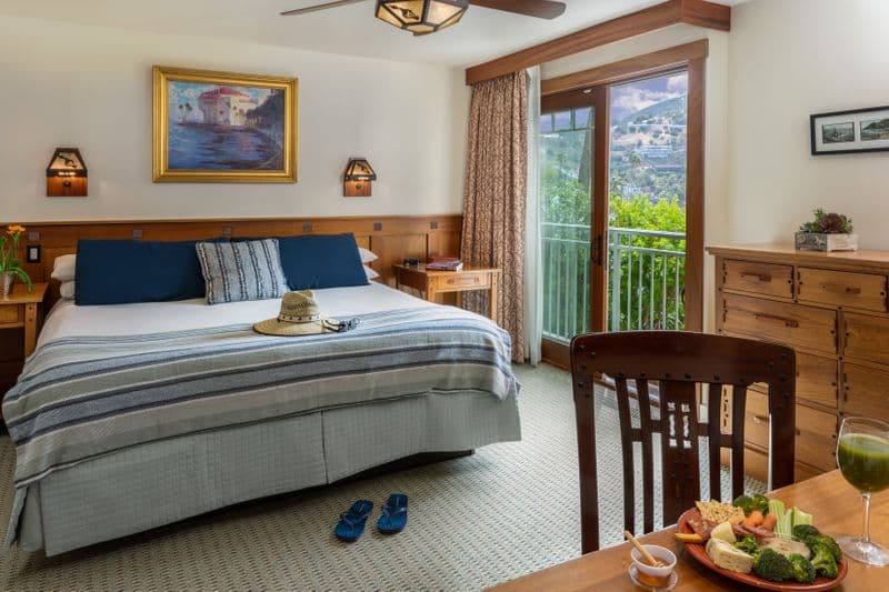 A cozy hotel bedroom featuring a neatly made bed with navy blue and striped accent pillows, a wooden dresser, and an open sliding glass door leading to a private balcony with a view of a lush palm tree and a neighboring hillside.